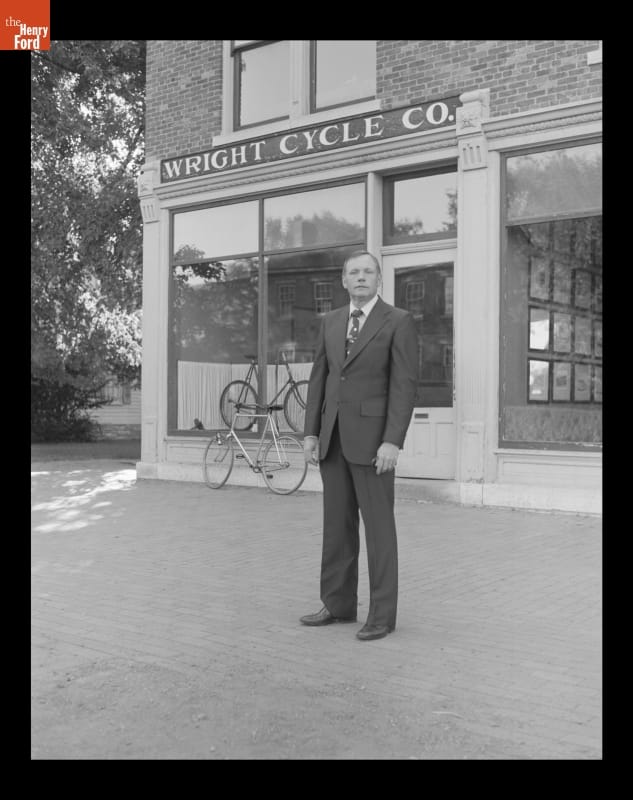 Man in a suit stands before a brick building with a bicycle outside and one in the window