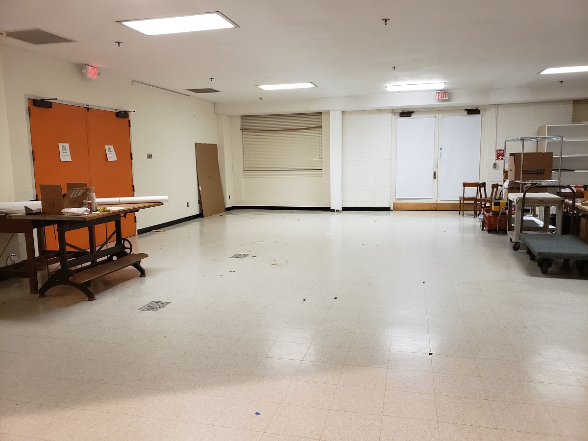 An image facing the doors and a very empty corner of the old Photo Studio Room with orange double doors on one wall and white double doors on another, with table on one side and other items in a clump on the other