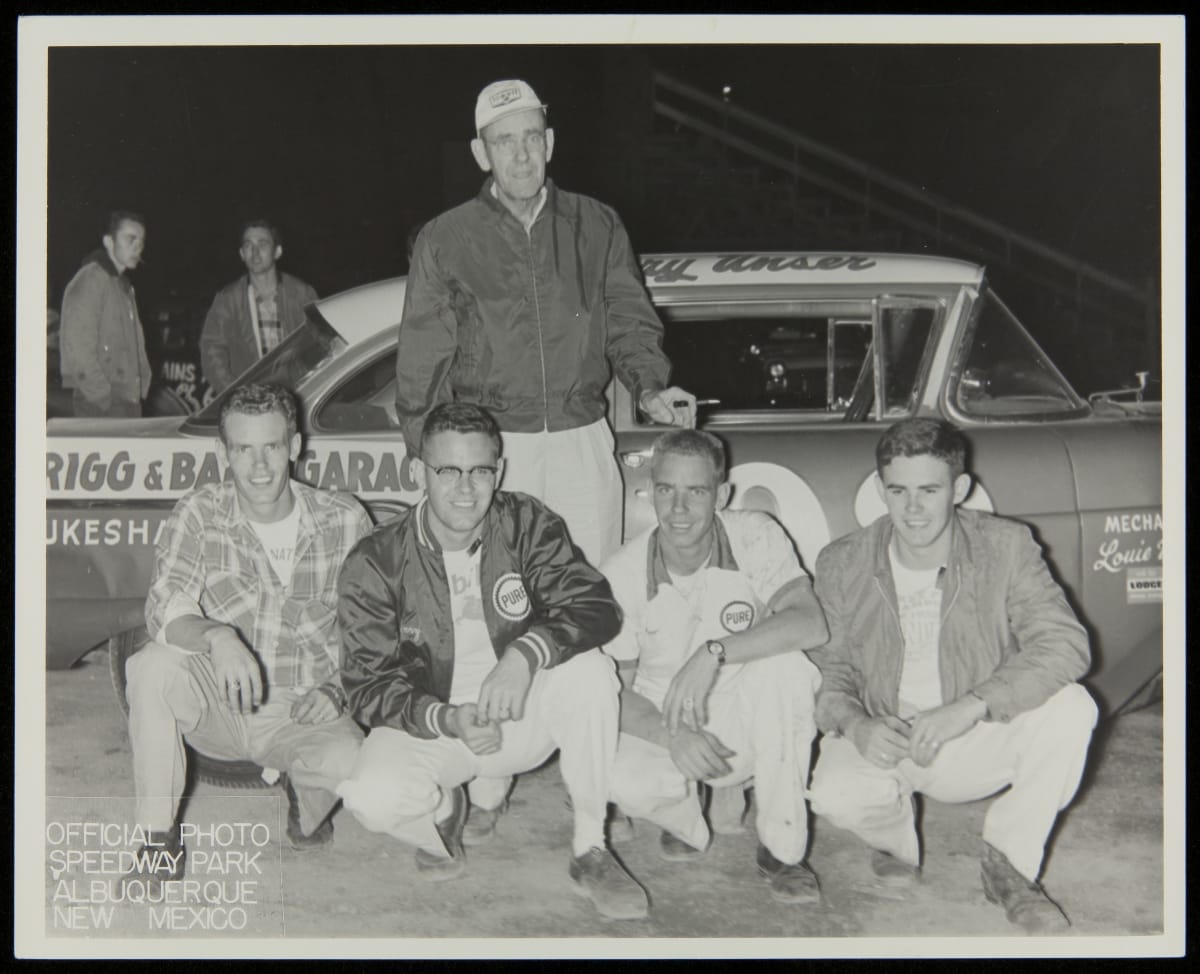 Unser Family at Speedway Park, Albuquerque, New Mexico, 1950-1959 Black-and-white photo of man standing in front of racecar, with four younger men kneeling in front of him