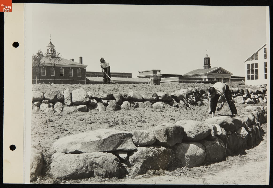 Cultivating and Planting Activity at Soybean Laboratory, Greenfield Village, Dearborn, Michigan, 1937-1950 Two young men or boys work in tiered garden plots divided by boulders with buildings in the background