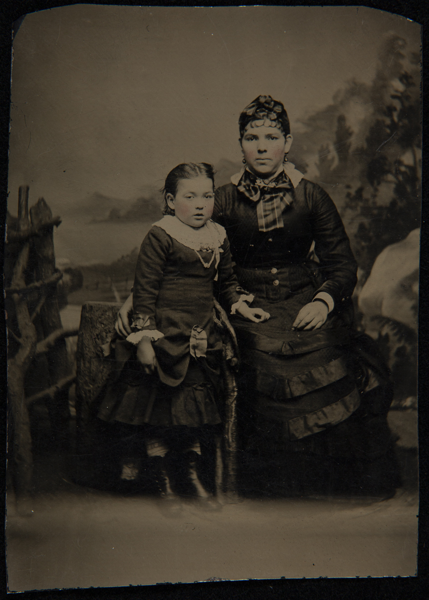 Seated woman and standing little girl, both in elaborate dresses, in front of a painted backdrop with logs as props