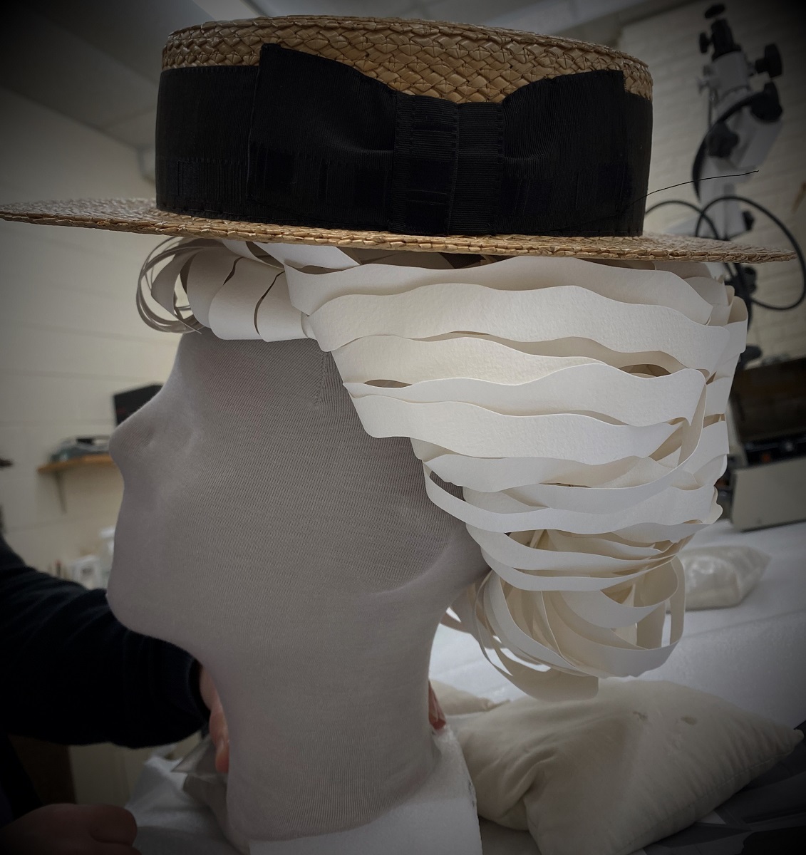 Gray mannequin head with a white, cut-paper wig topped with a straw boater hat with wide black ribbon