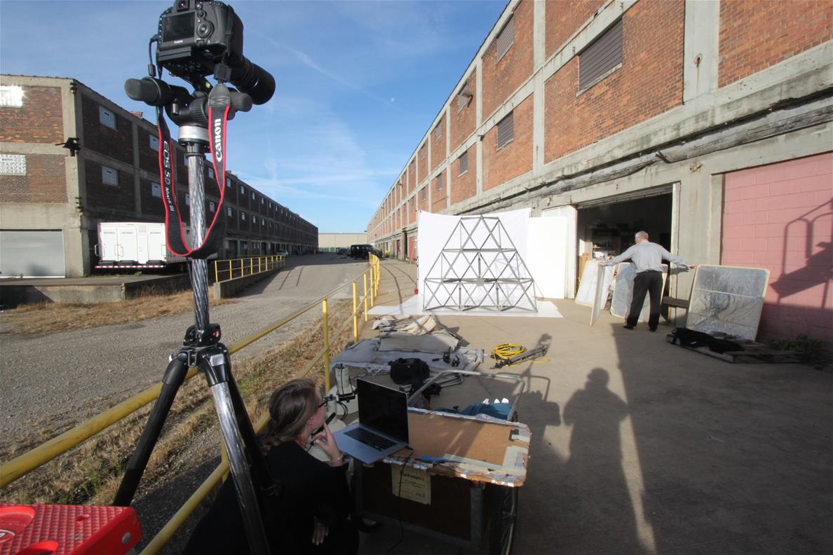 Camera on tripod in foreground pointed toward a large metal truss in front of white background, with large, low brick buildings extending into the distance on both sides