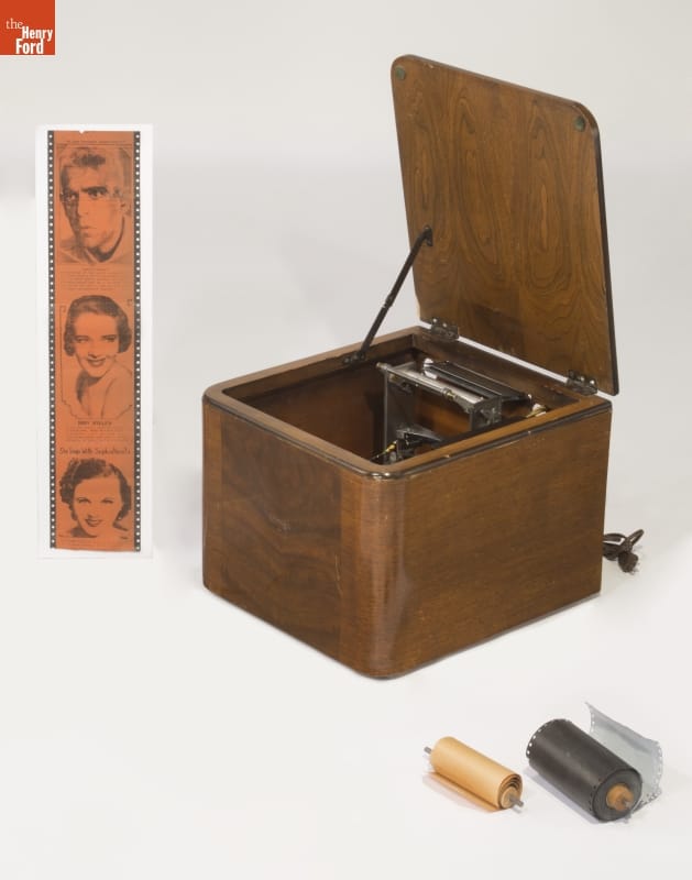 Wooden box with machinery visible inside; two rolls in foreground of picture and strip with images of several faces on left side of image