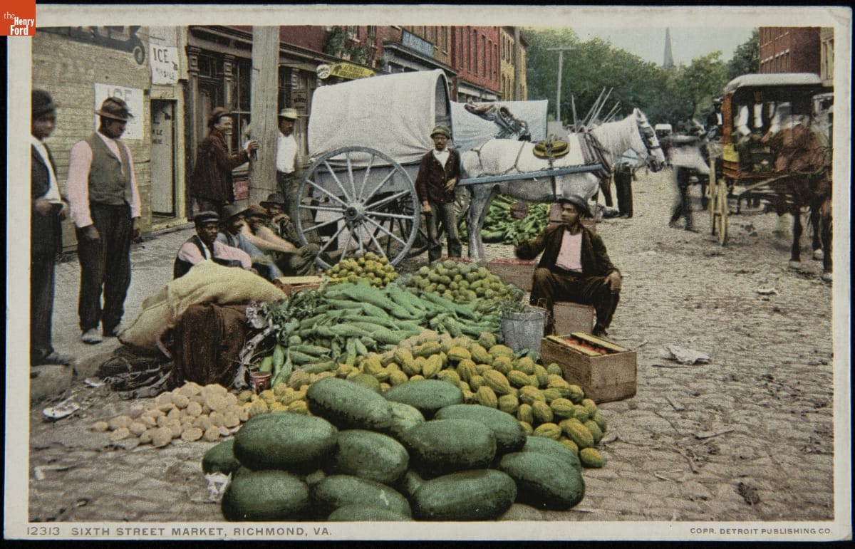 Men of color sit and stand around a piles of different vegetables at one side of a cobblestone street with buildings and horses and carriages behind them