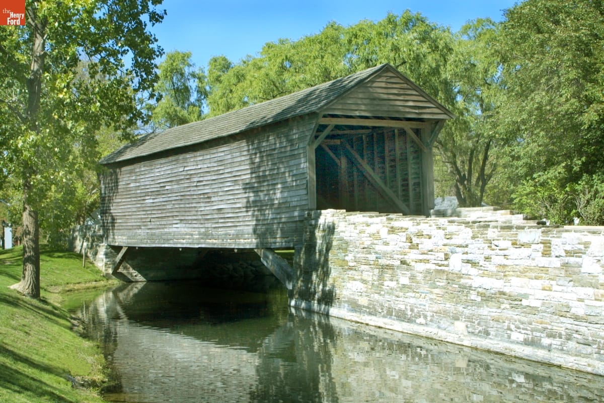 A wooden covered bridge crosses a stream with a stone wall on one side and trees behind it