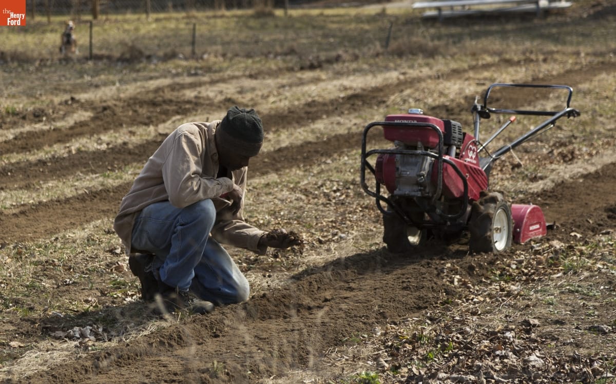 A man kneels in a field by a piece of equipment, looking at a handful of dirt he is holding 