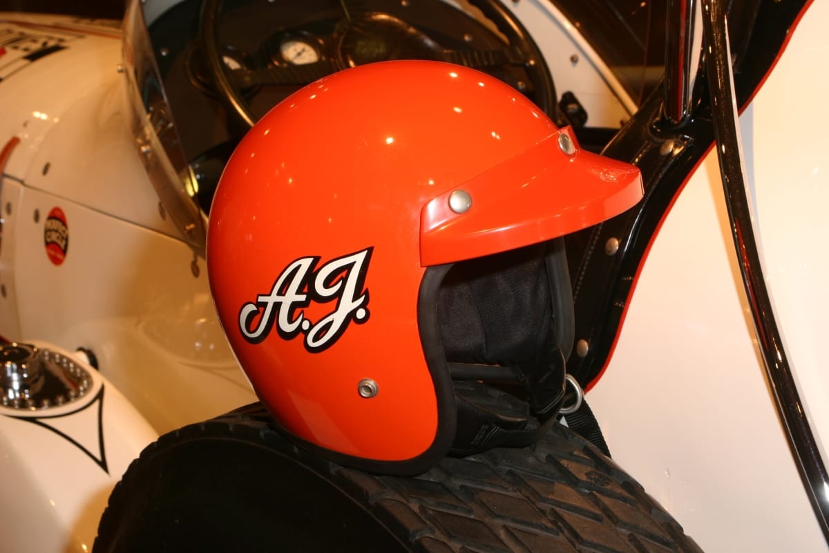 Orange helmet with white initials "A.J." on side, sitting on wheel of white race car
