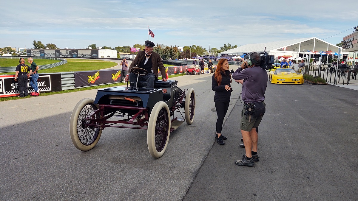 Man sits in boxy open early car on racetrack; a woman stands nearby being filmed by a cameraman