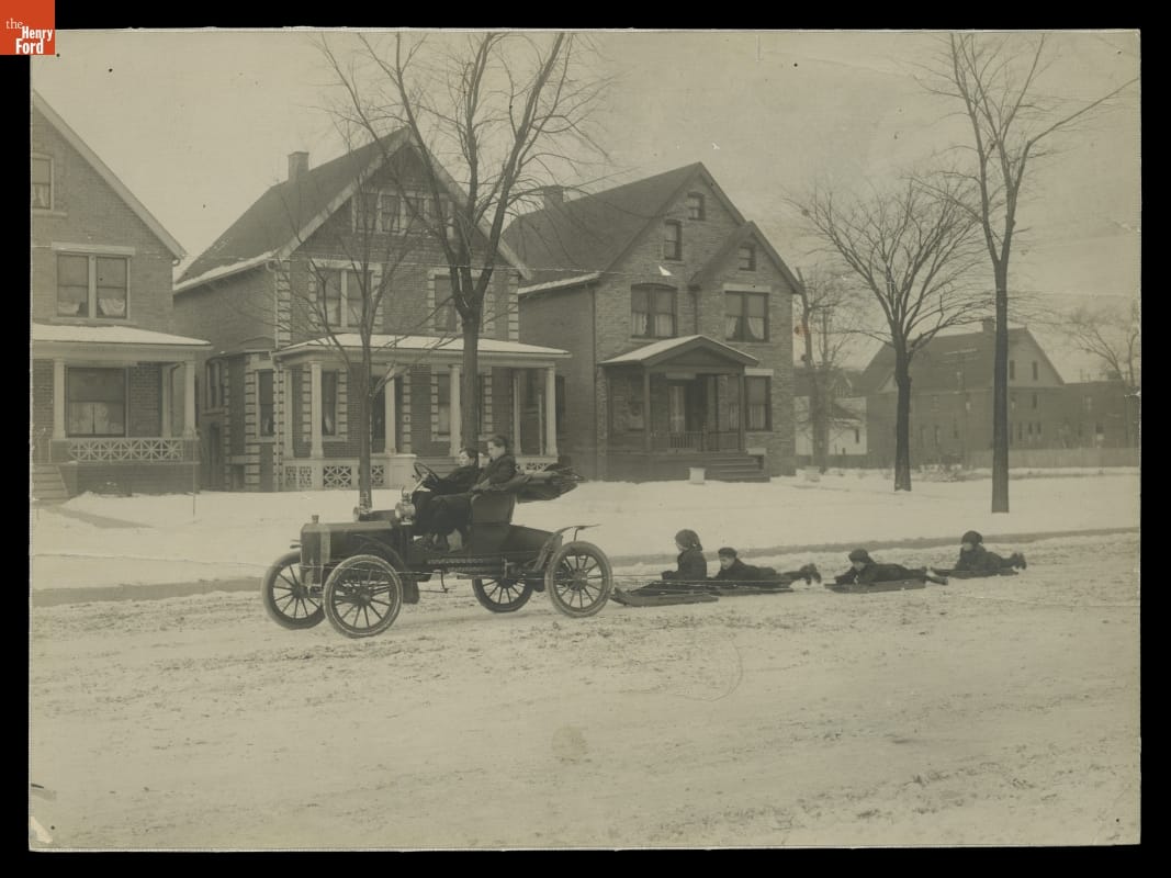 Car with two people in it, driving down snowy street with four children on sleds tied to bumper behind; trees and houses in background