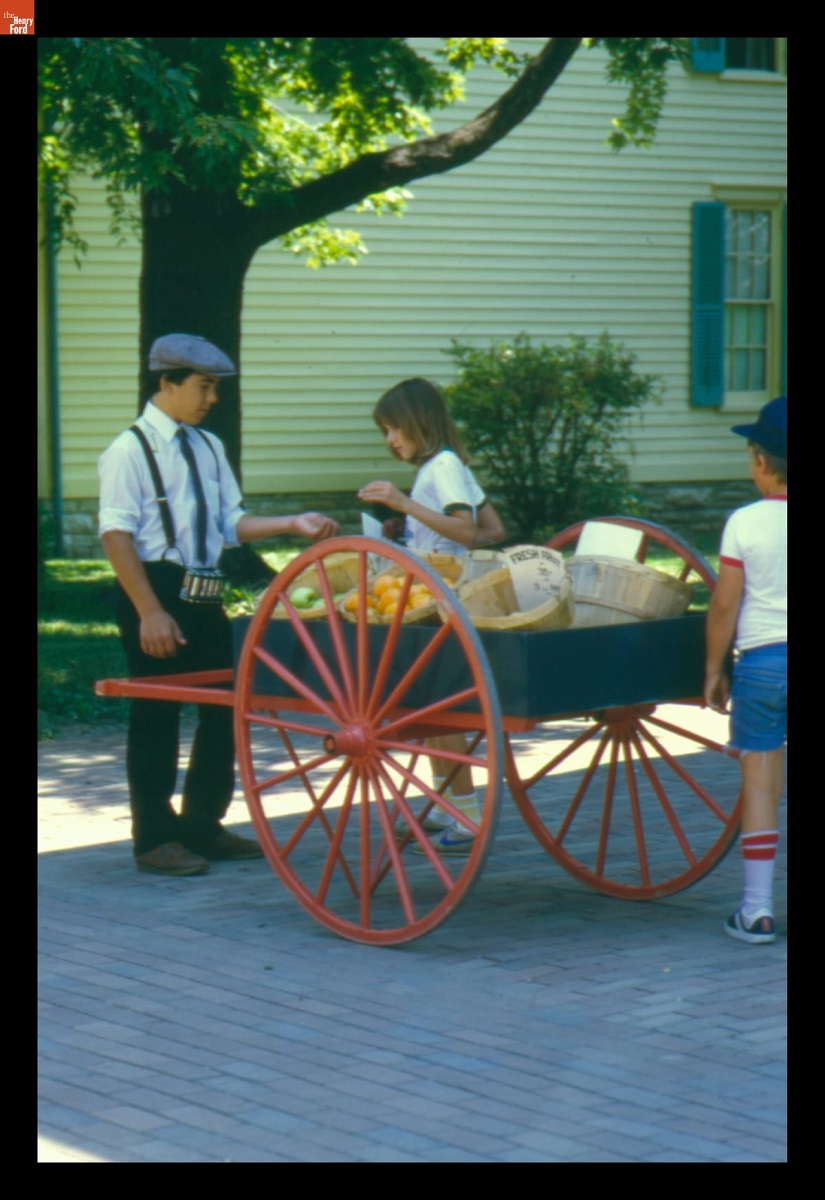 Man in suspenders and cap stands in front of cart filled with baskets, with two children nearby