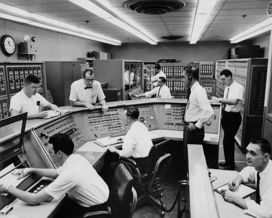 Group of men sitting and standing at computer consoles and banks