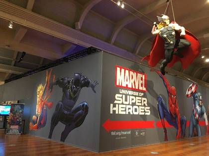 Entrance to the "Marvel: Universe of Super Heroes" exhibition in Henry Ford Museum of American Innovation Gray wall with images and text in larger building; hanging figure of superhero in right foreground