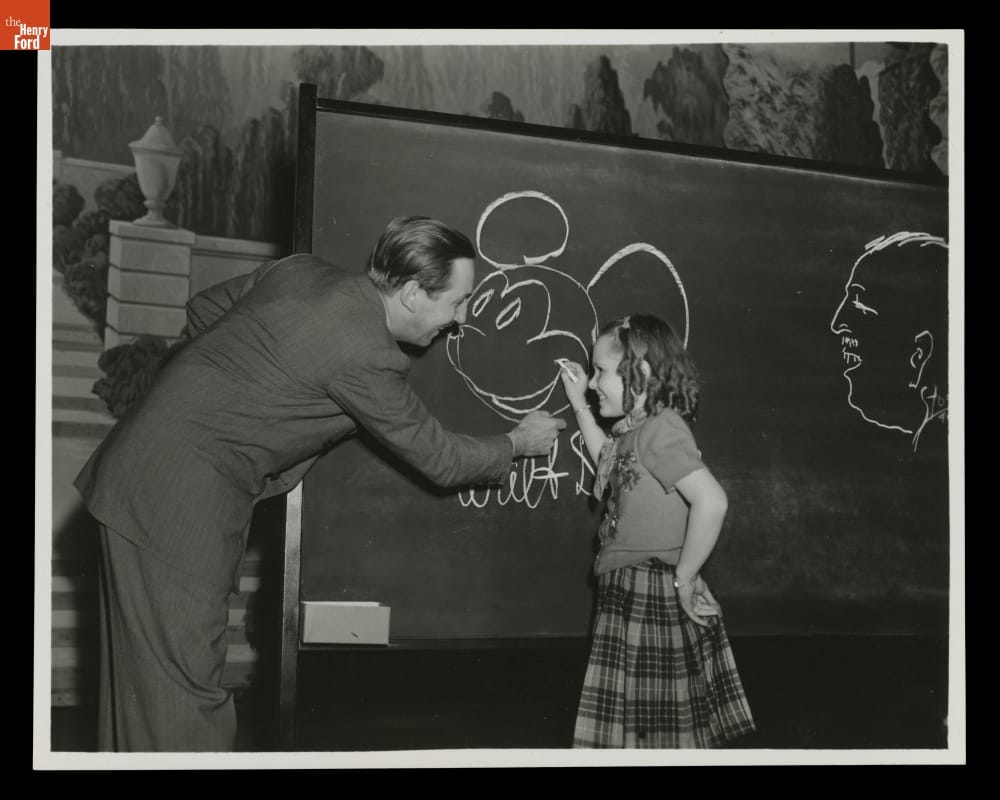 Man in suit and young girl draw Mickey Mouse's head on a chalkboard