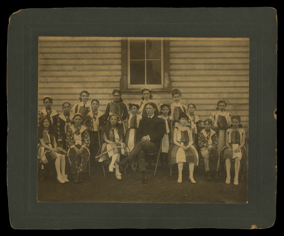 Students Dressed in Patriotic Costumes, with Teacher, circa 1905 Matted black-and-white photo of group of children wearing stars and stripes sitting and standing with a man in a suit in front of a wooden building