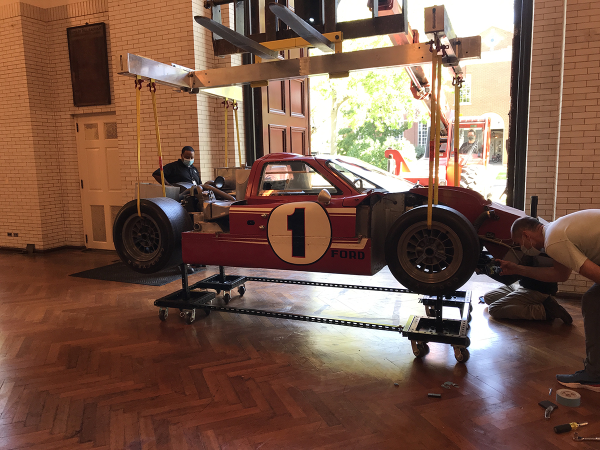 Red race car with part of body removed and large "1" on side, suspended from a crane-like gantry in front of a large door; one person stands nearby and another examines the car