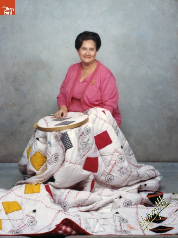 Woman with pink outfit and dark hair sits with a quilt over her, part of which is in a needlework frame on her lap