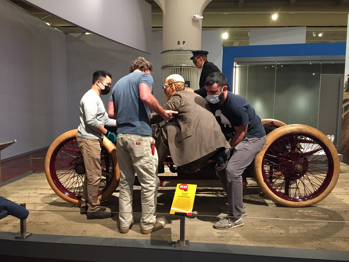 Three men move a mannequin into place on the running board of a car; another mannequin is inside the car
