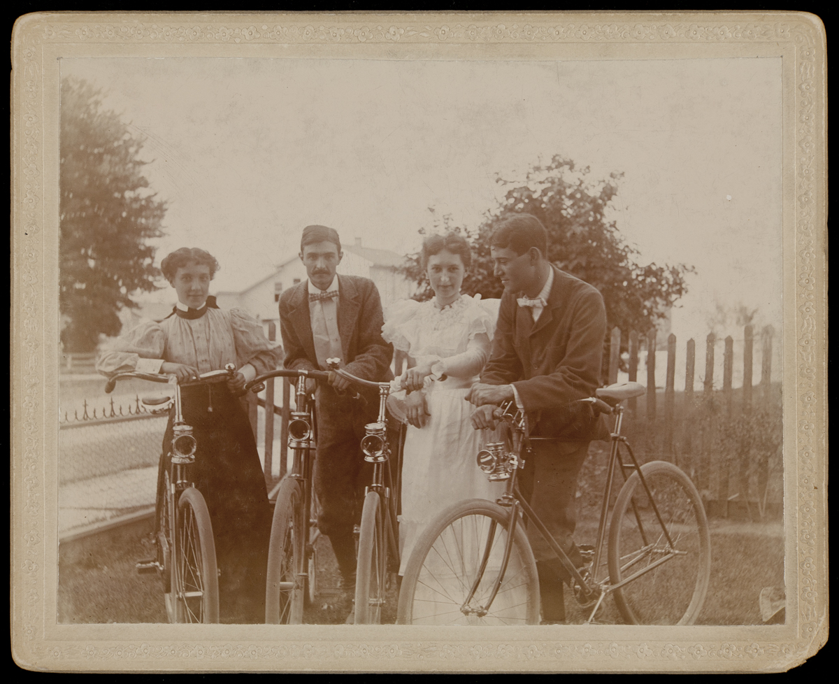 Two women in dresses and two men in suits, each standing next to a bicycle