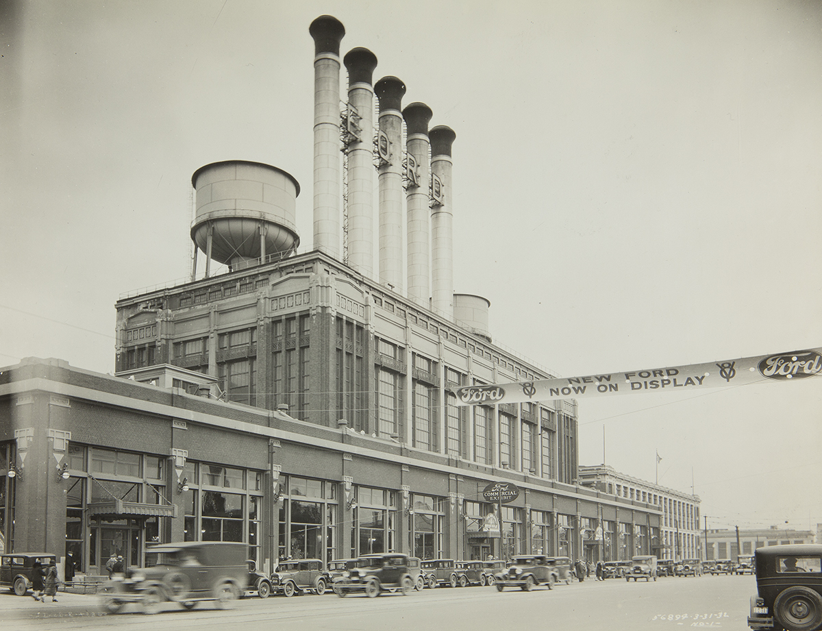 Factory topped with several smokestacks and a water tower, with cars driving by in front