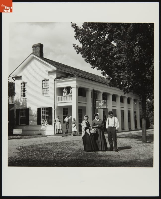 Black-and-white photo of people standing outside and on the first- and second-story balconies of a long wooden building with many columns 
