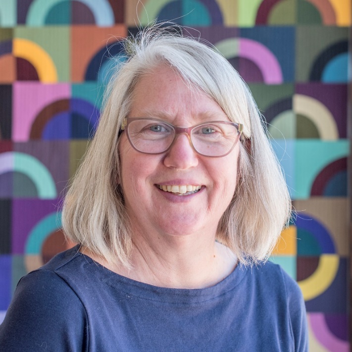 Light-skinned woman with blonde hair and glasses, wearing blue top, in front of a colorful backdrop