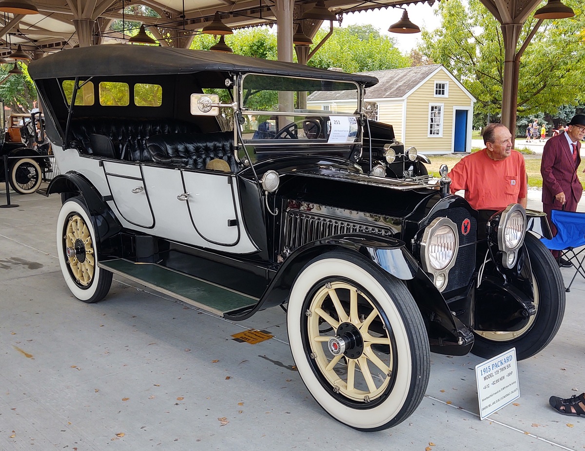 1915 Packard Twin Six at Old Car Festival 2022 Old-fashioned car with white doors and black top, with open space where windows and dash would be, displayed in an open building with people nearby