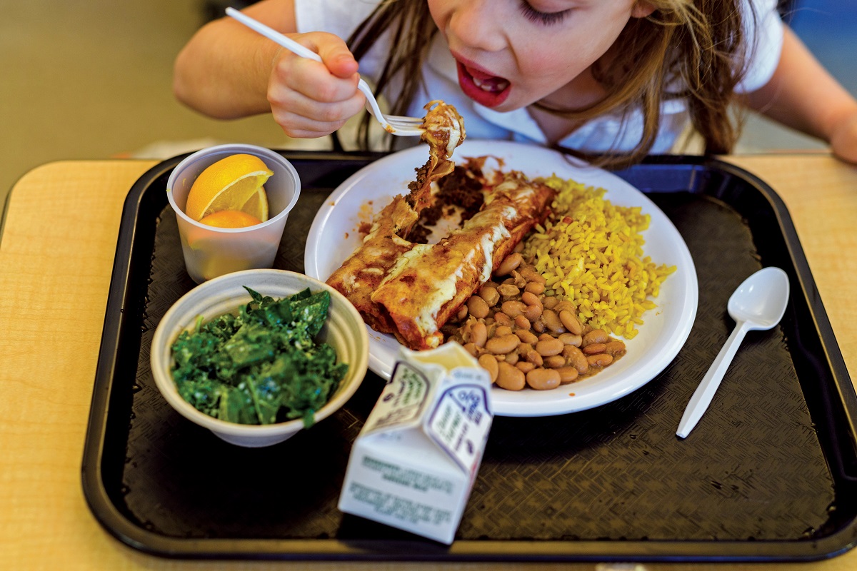 A student in New London, Connecticut, tries enchiladas A child lifts a fork of food to her mouth behind a cafeteria tray holding enchiladas, beans, rice, orange sections, a salad, and a milk carton