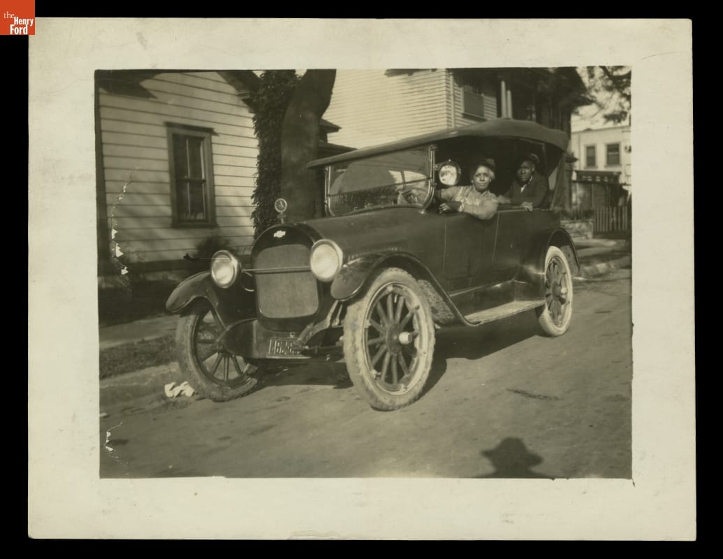 Woman at wheel of old-fashioned car with man visible in seat behind her; houses behind them