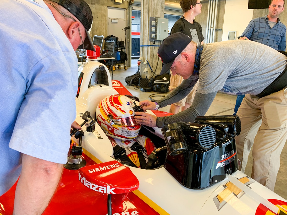 One person adjusts a camera on the helmet of another person sitting in an open cockpit race car; additional people stand nearby