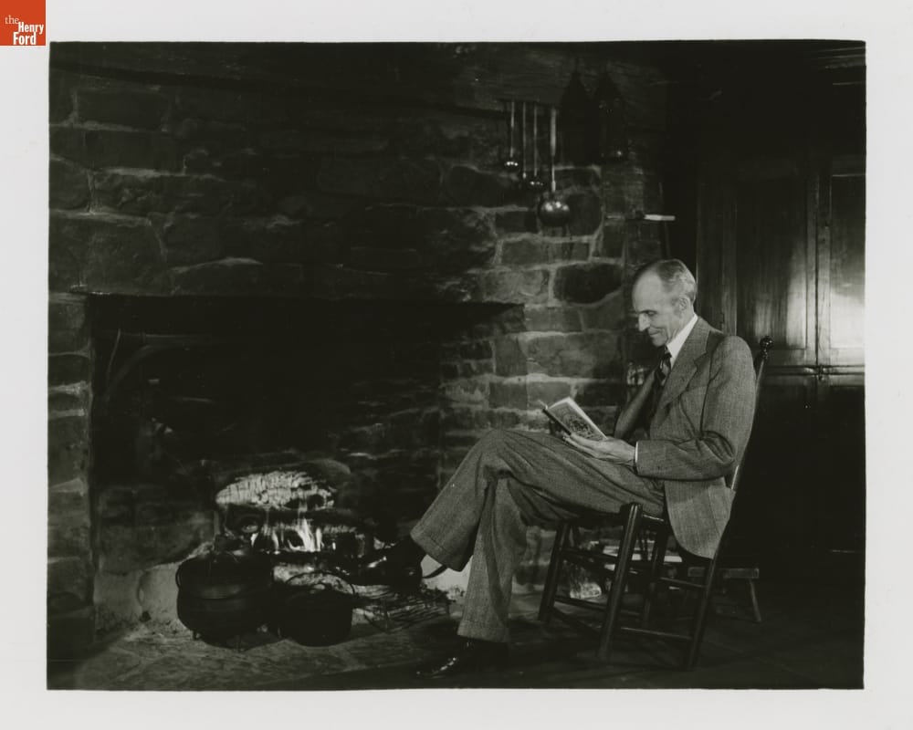 Man wearing suit sits in a chair by a fireplace, reading a book