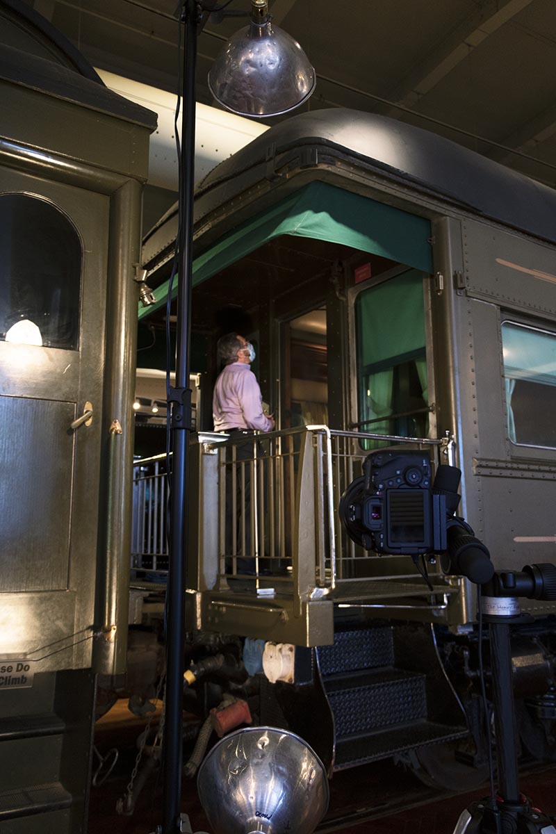 Man standing on back platform of railcar in large room, with camera and light in foreground