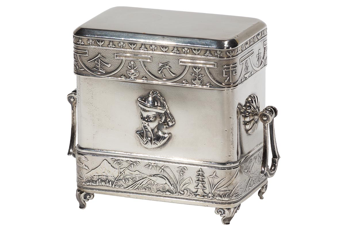 Silver box on small legs, with handles on either side and relief on the front of man's face and torso with long drooping mustache and wearing a hat