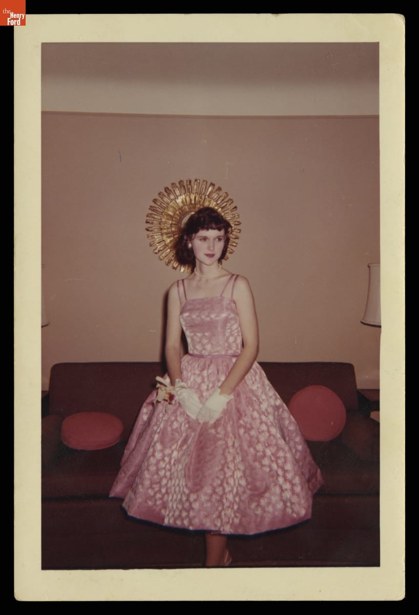 Woman in pink sleeveless dress with full skirt, wearing white gloves, poses in front of a couch