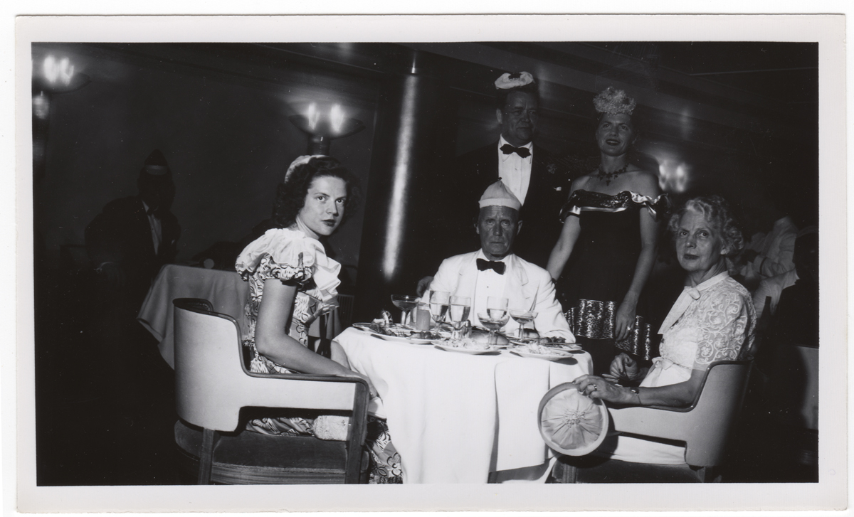 Black-and-white photo of three people seated at a table look toward the camera; two more people stand behind the table