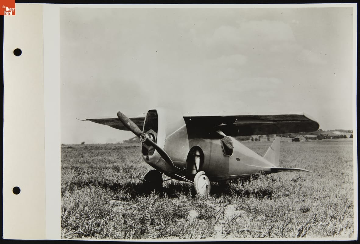 Black-and-white photo of small airplane parked in field with trees and buildings in the distance behind it