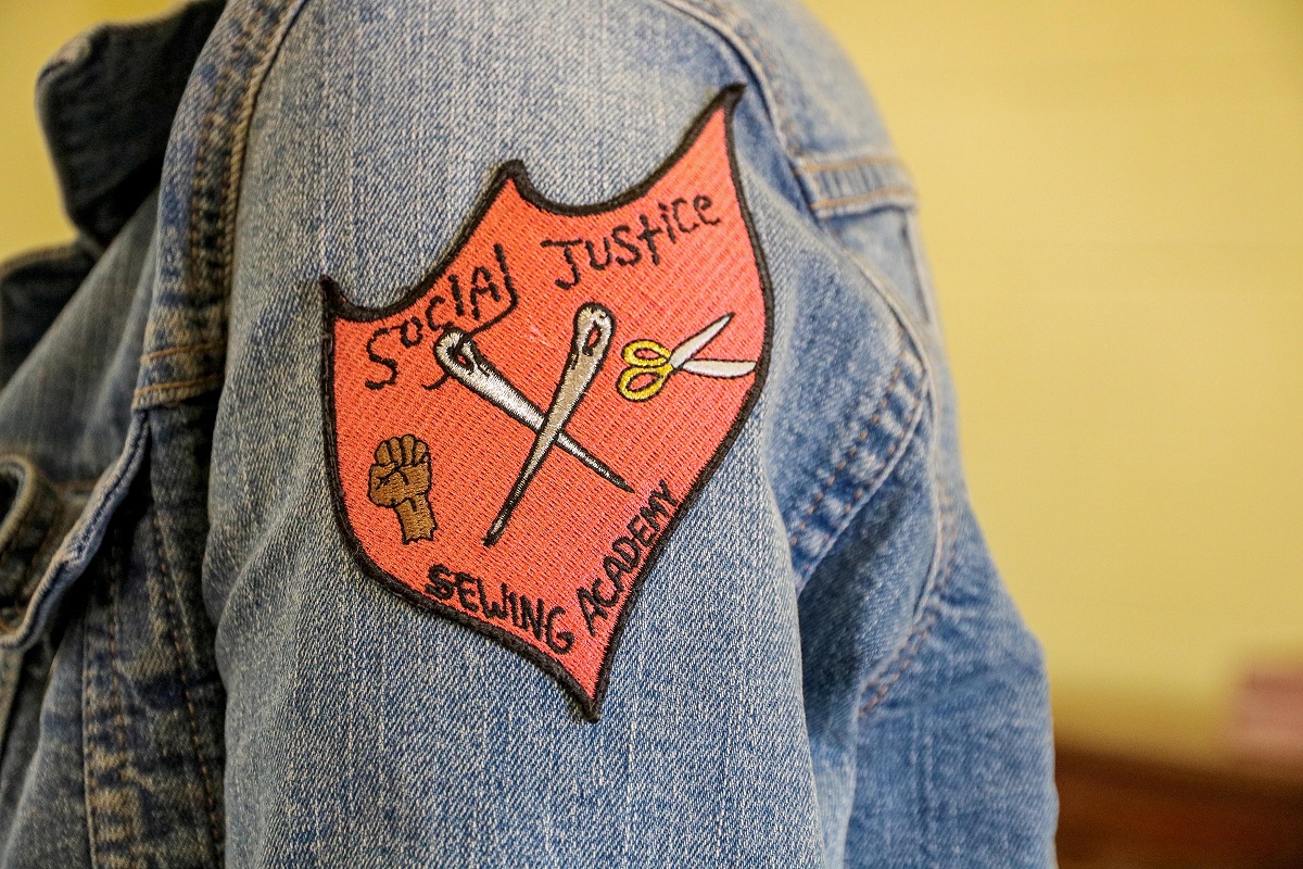 Red, shield-shaped patch with text and images of two crossed sewing needles, an upraised brown fist, and a pair of scissors, on the sleeve of a jean jacket 