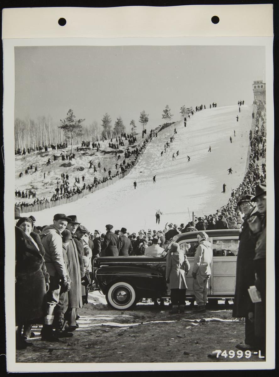 People on steep, snow-covered ski slope, with crowds on either side and more crowds and a car in the foreground