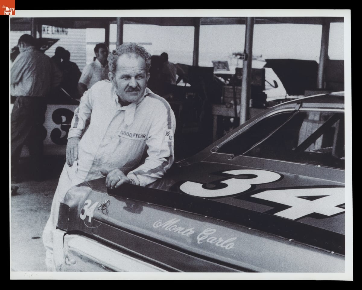 Wendell Scott with His 1971 Chevrolet Monte Carlo at the World 600, Charlotte Motor Speedway, May 24, 1972 Man in jumpsuit with short curly hair and mustache leans against back of race car, with other race cars, people, and equipment in the background
