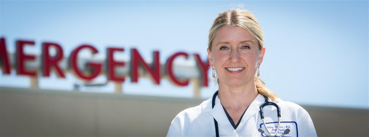 Hillary Goodwin, Auburn, California Light-skinned blonde woman in medical coat with stethoscope around neck stands in front of sign reading "EMERGENCY"