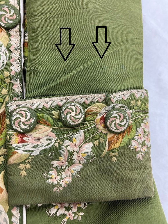 Detail of moss-green coat sleeve with elaborate embroidery and embroidery-covered buttons; two arrows point to two slight picks in the fabric above the cuff