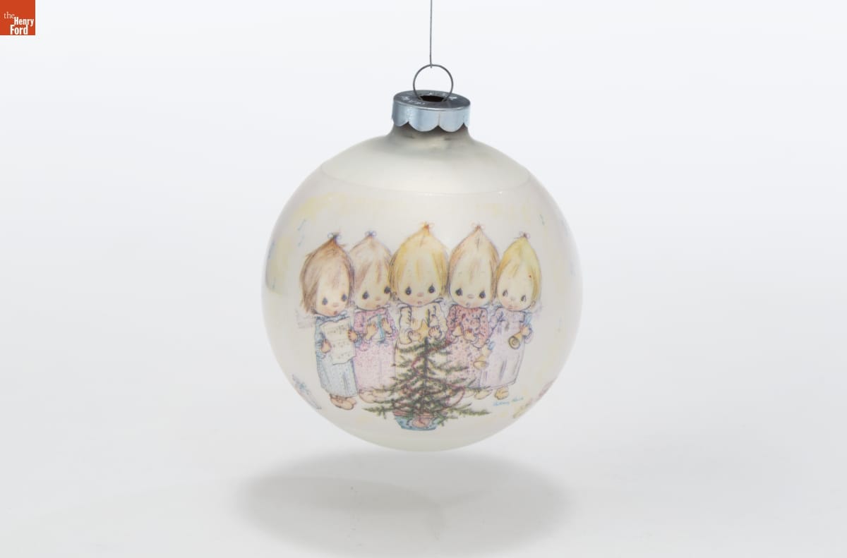 Round white Christmas ornament with image of 5 young children with fine hair holding musical instruments and sheet music around a small Christmas tree