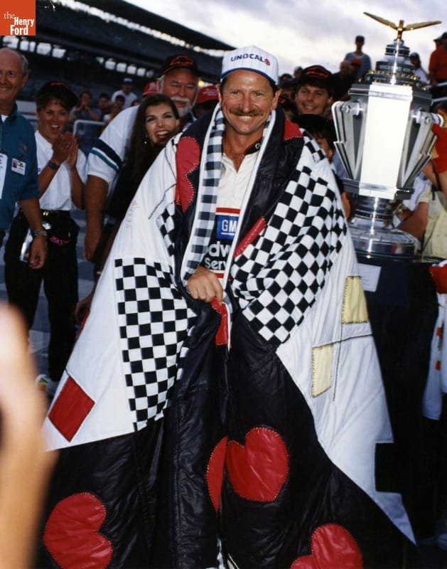 Man wrapped in quilt wearing baseball cap stands among other people with a large trophy and grandstands in the background