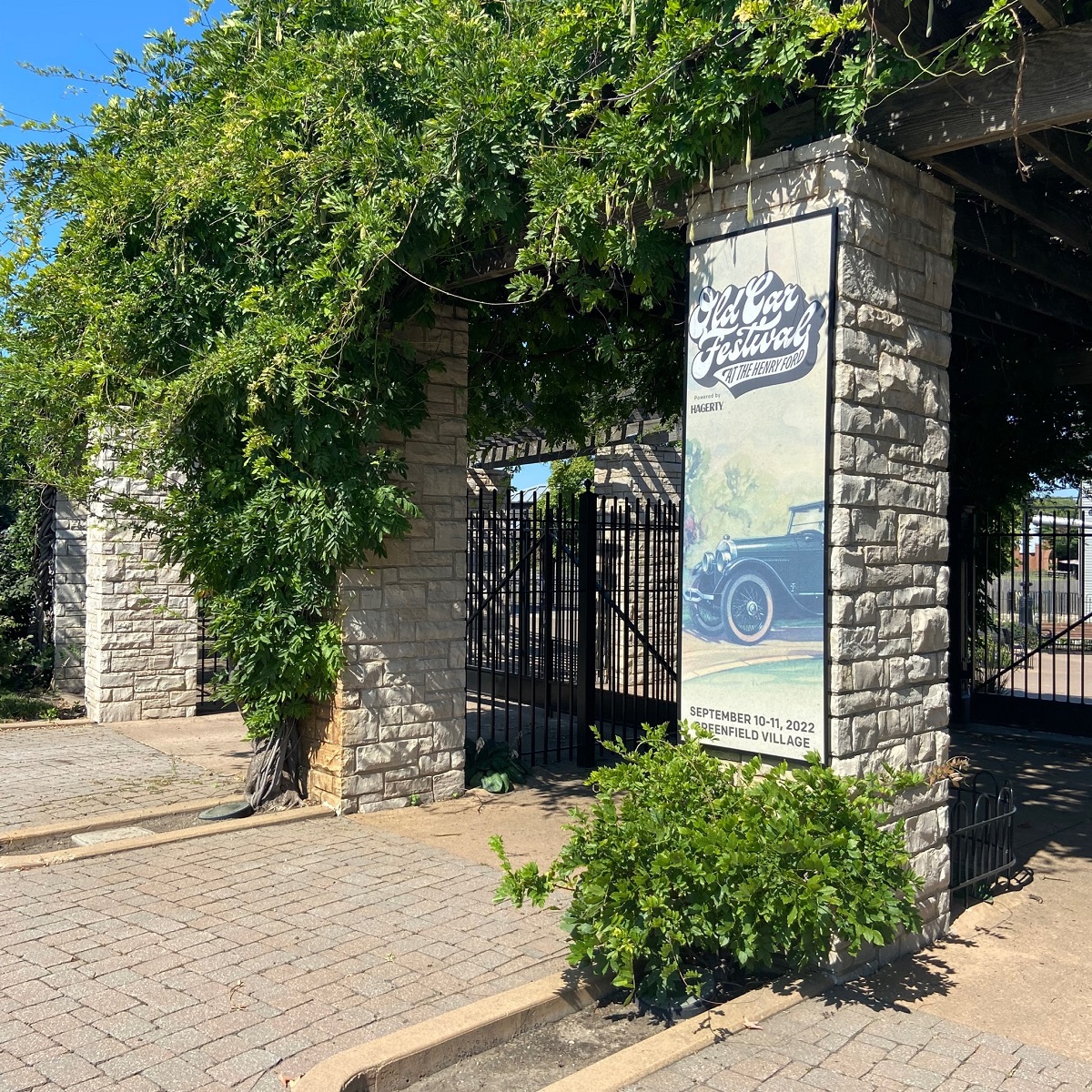Old Car Festival 2022 banner by the Greenfield Village entrance gates Large stone columns with black iron gates between them and green vines growing down over the tops; one is hung with a banner with text and an image of a car