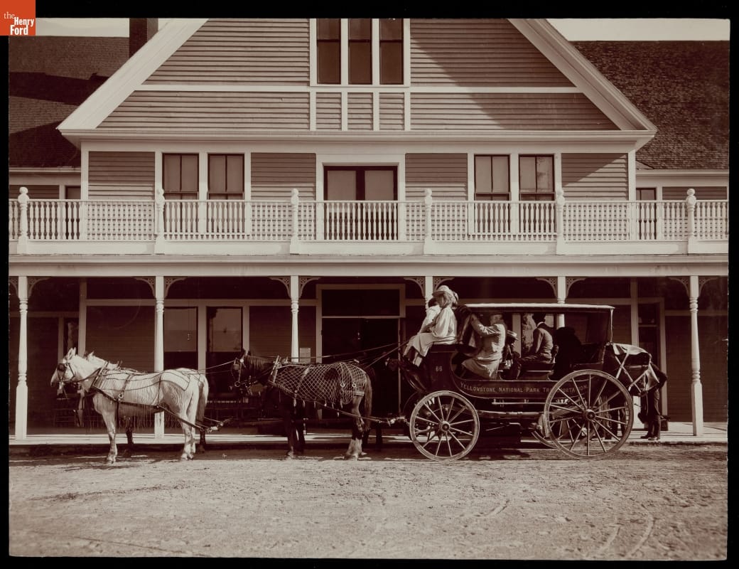 Black-and-white photo of a horse-drawn stagecoach filled with passengers in front of a building