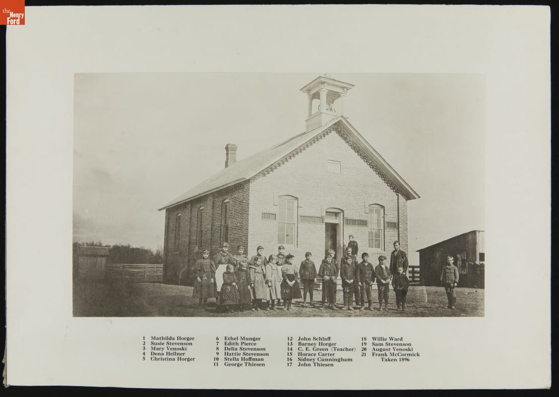 Group of children pose outside of a small brick building; also contains text key with the names of those in the photo