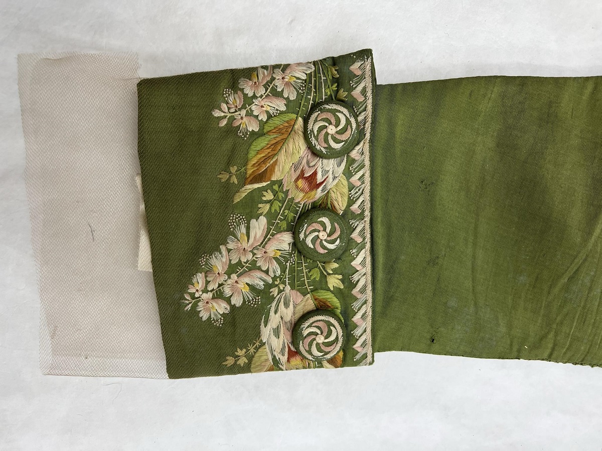 Detail of moss-green coat sleeve with detailed floral embroidery on cuff; tulle extends from end of cuff