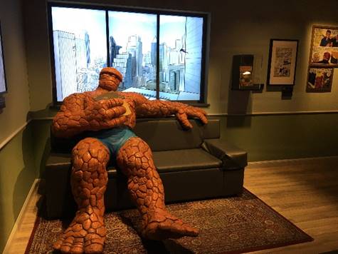 Photo op with The Thing in "Marvel: Universe of Super Heroes" exhibit Oversize mannequin with orange reptilian skin sitting on couch with illuminated drawing of city skyline behind him; additional cases and framed drawings on wall to right