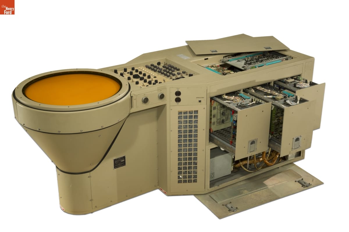 Large piece of beige metal equipment with round yellow screen (?) at one end and buttons and switches; open panels on top and sides showing complex wiring