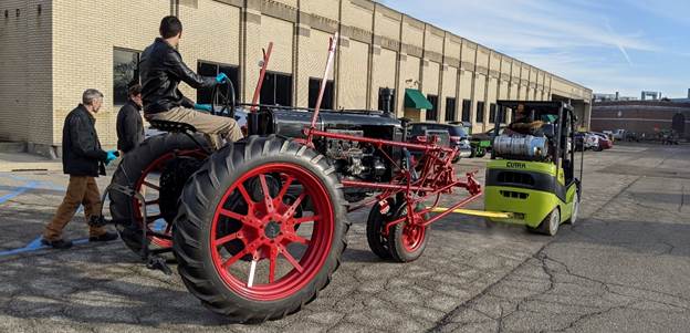 Small vehicle towing a large gray tractor with red wheels, with a man sitting on the tractor and a couple others walking next to it